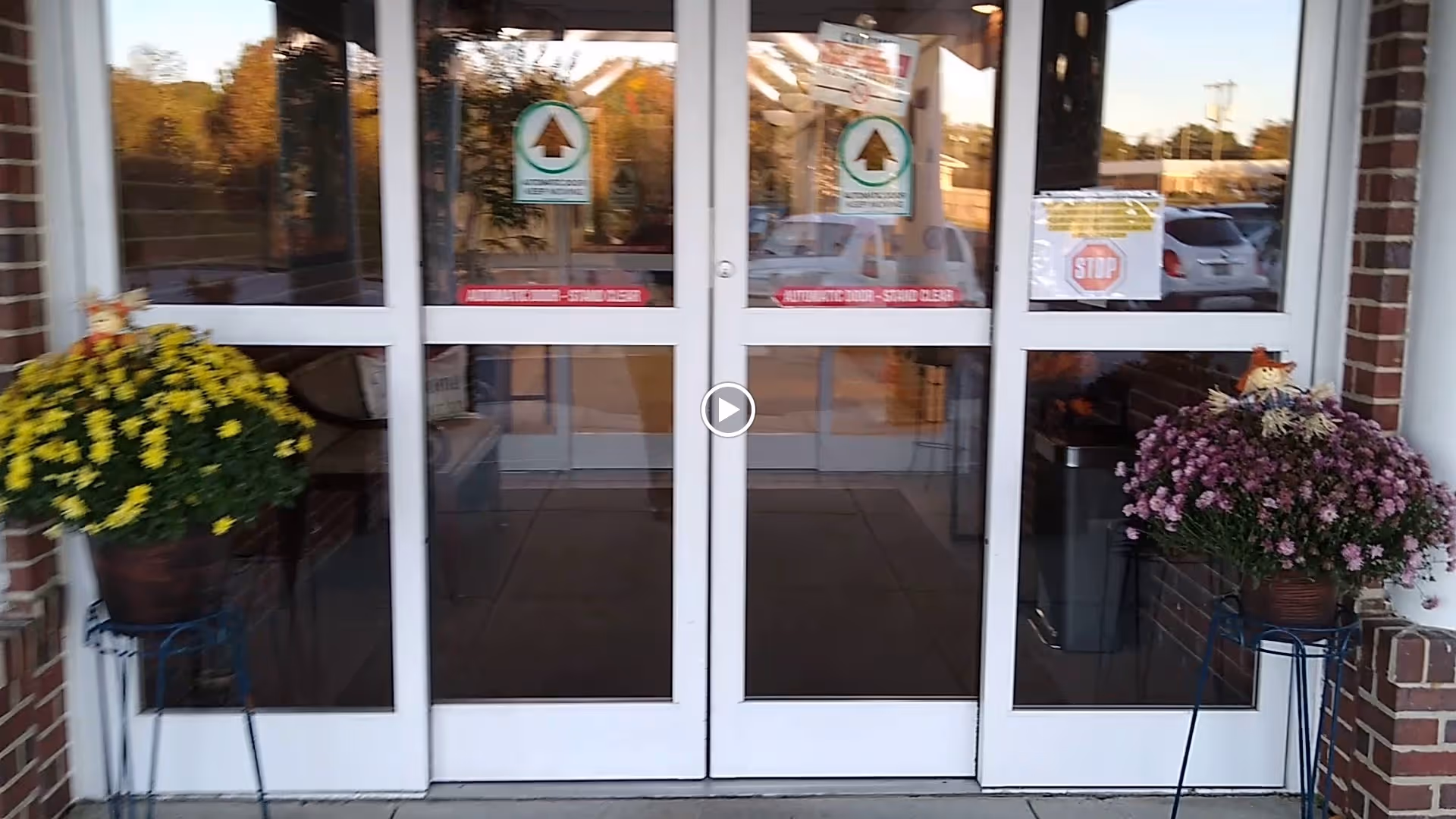 Glass double doors entrance to a building with white frames. On either side of the doors are large flower pots on stands, one with yellow flowers and the other with purple flowers. There are signs on the doors including a stop sign and notices about the automatic doors. The building exterior is brick and there are cars visible in the parking lot outside.