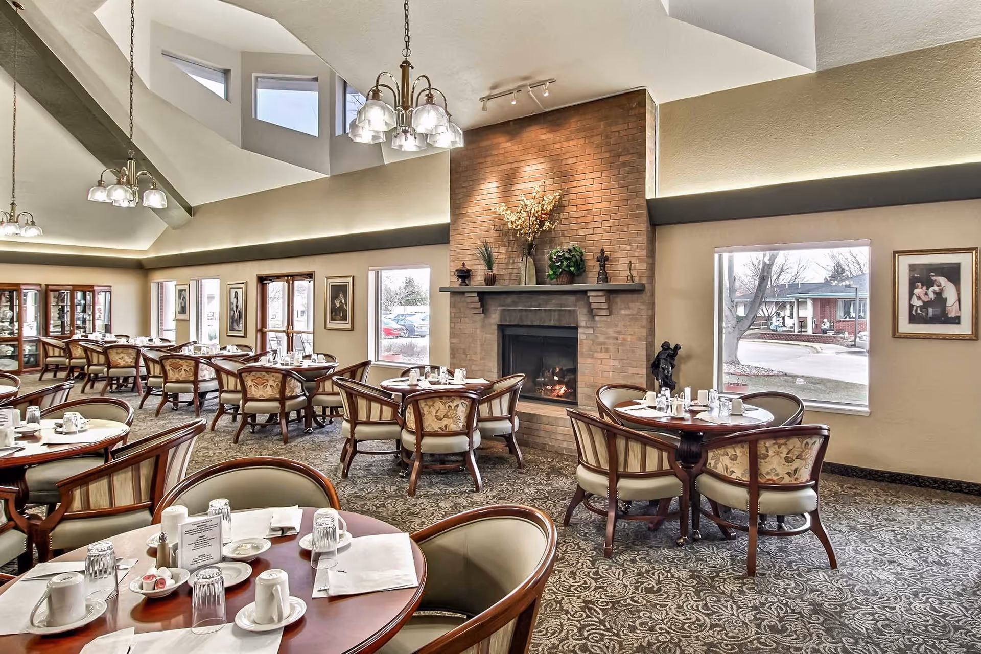 A spacious dining room with multiple round tables set with white cups, plates, and napkins. The room features a large brick fireplace with decorative plants on the mantel, patterned carpet, and large windows letting in natural light. There are framed pictures on the walls and elegant chandeliers hanging from the ceiling.