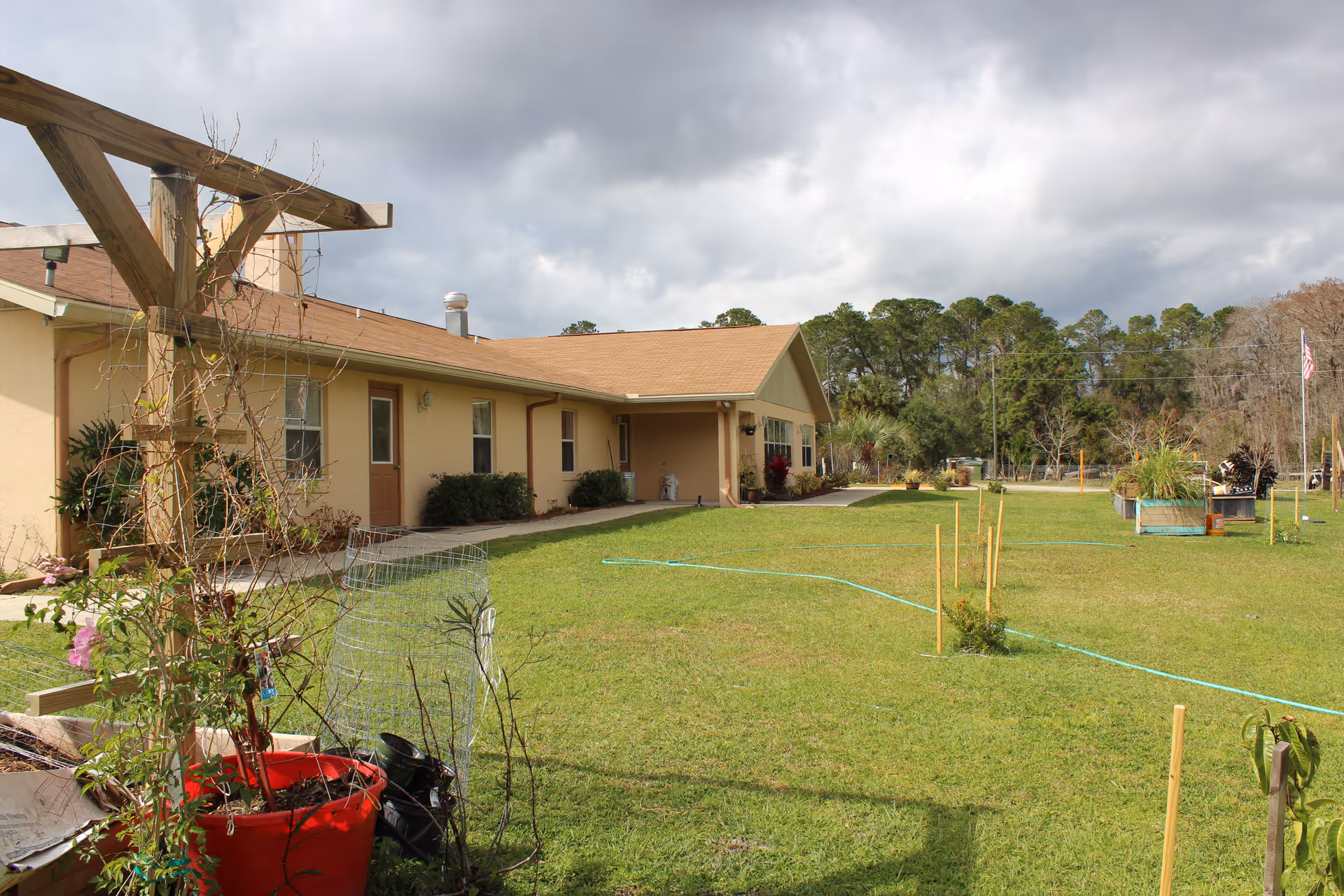 Single-story beige building with a wide grassy lawn, garden planters and a trellis under a cloudy sky.