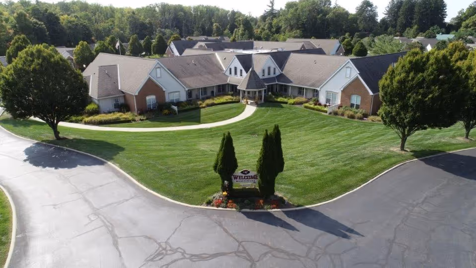 Aerial view of Wedgewood Estates senior living facility showing a large, single-story building with multiple peaked roofs surrounded by well-maintained green lawns, trees, and a curved driveway. There is a small landscaped area with a welcome sign at the center front of the building.