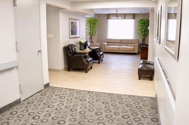 A bright and clean senior living facility common area with a beige sofa at the far end, two dark leather armchairs around a small round table with a flower vase, two tall green plants on either side of the sofa, and a patterned carpet leading into a light wood floor area.