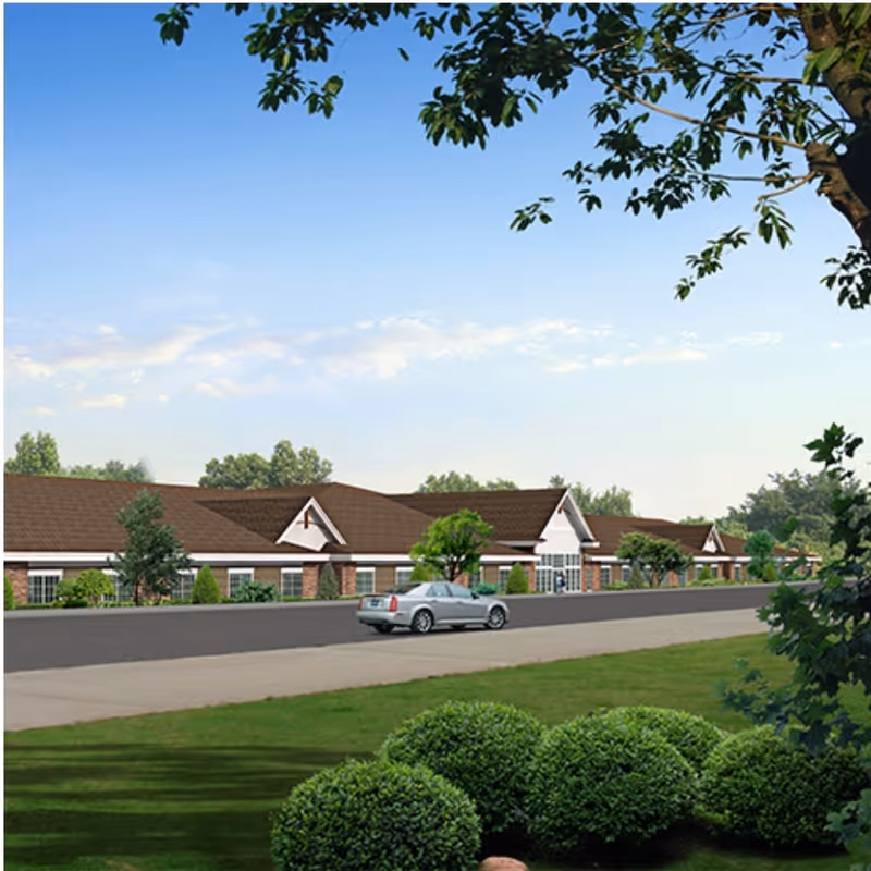 Exterior view of a single-story senior living facility building with a brown roof, white walls, and multiple windows. A silver car is parked on the road in front of the building. The foreground features green bushes and a grassy area, with trees framing the top and right edges of the image under a clear blue sky.