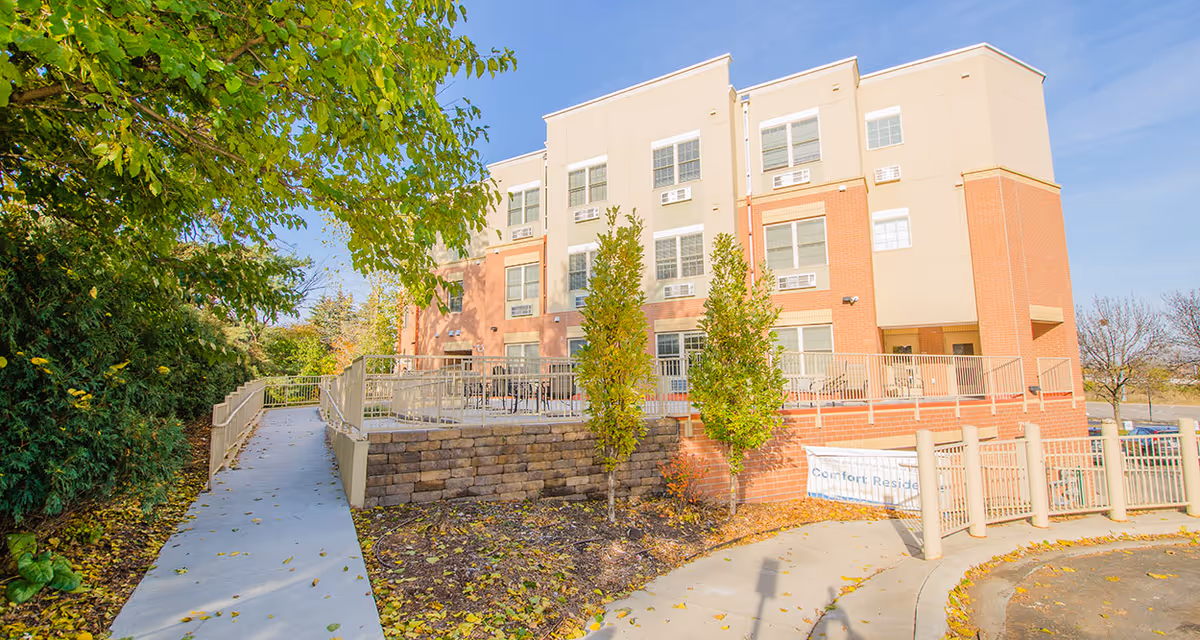 Exterior front of a multi-story senior living building with a paved walkway, raised patio with railings, and landscaped trees under a clear sky.