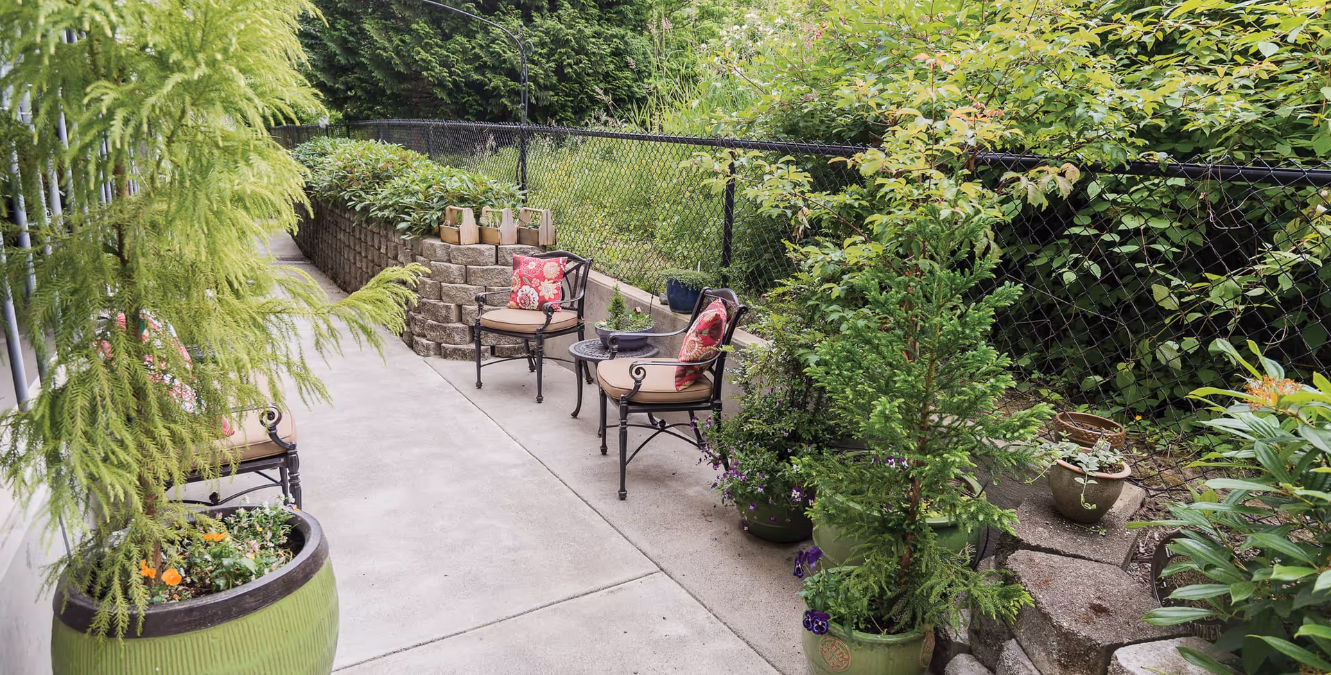 Outdoor patio area with concrete walkway, surrounded by various green plants and shrubs in pots and garden beds. There are two metal chairs with beige cushions and red patterned pillows, and a small round table between them. A black chain-link fence runs along the right side of the walkway.