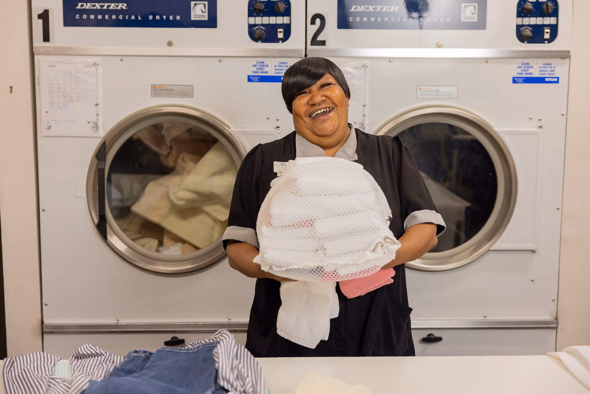 A smiling woman holding a stack of folded laundry in front of two commercial dryers in a laundry room.