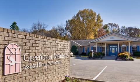 Brick sign reading 'Greenbrier Meadows assisted living by Americare' in the foreground with the facility entrance, parking area, and landscaping in the background.