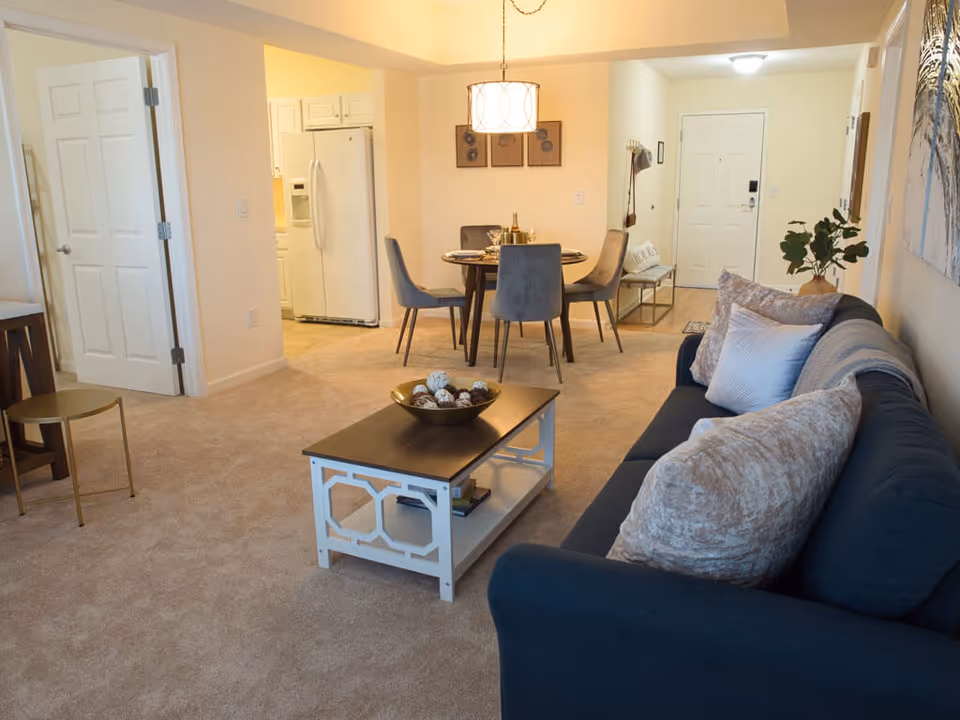 Open-plan living and dining area with a dark sofa and coffee table in the foreground and a dining table and kitchen refrigerator in the background.