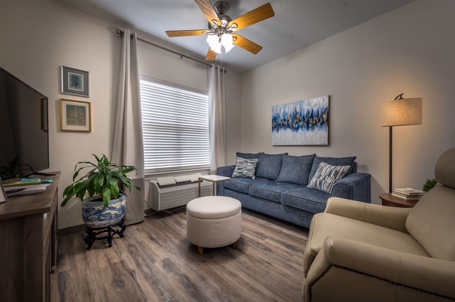 Cozy living room with a blue sofa, beige armchair, round ottoman, TV cabinet, potted plant, and wall art.