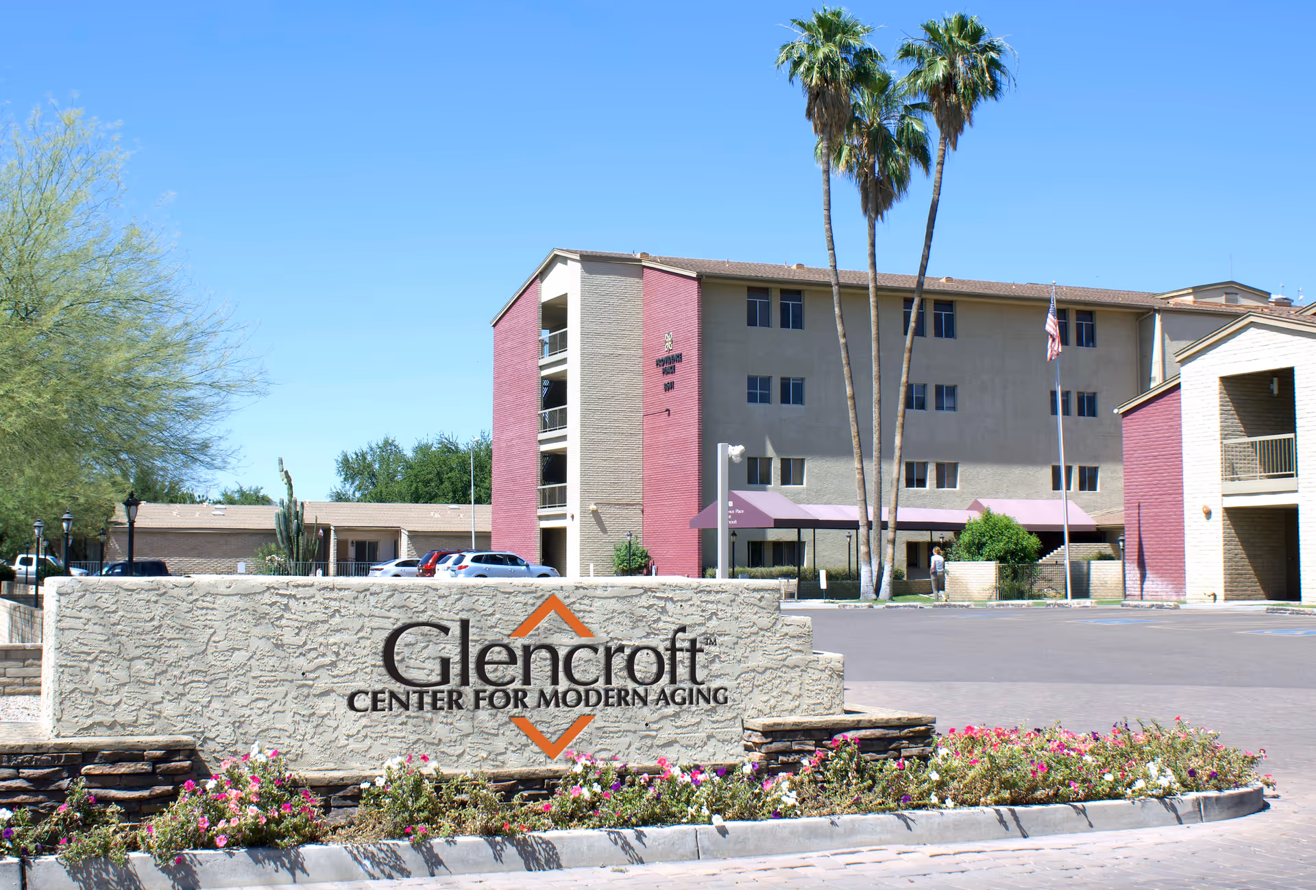 Exterior view of Glencroft Center for Modern Aging, showing a multi-story building with beige and reddish walls, palm trees, an American flag, parked cars, and a stone sign with the facility's name in front surrounded by flowers.