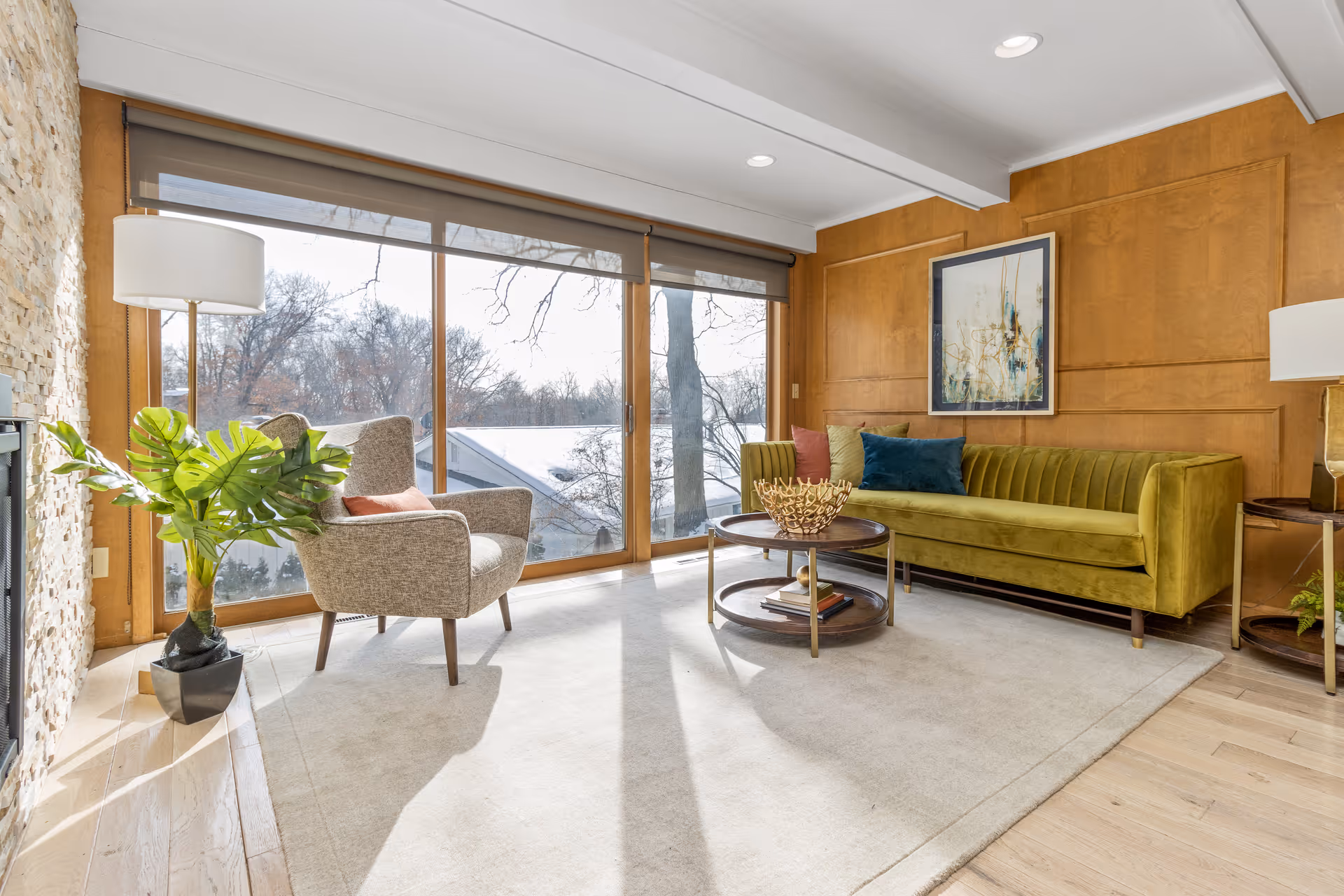 A bright living room with large floor-to-ceiling windows showing a view of trees outside. The room features a green velvet sofa with colorful cushions, a round wooden coffee table with decorative items, a gray upholstered armchair with a pink pillow, a tall floor lamp, a potted plant, and wood-paneled walls with a framed abstract painting.