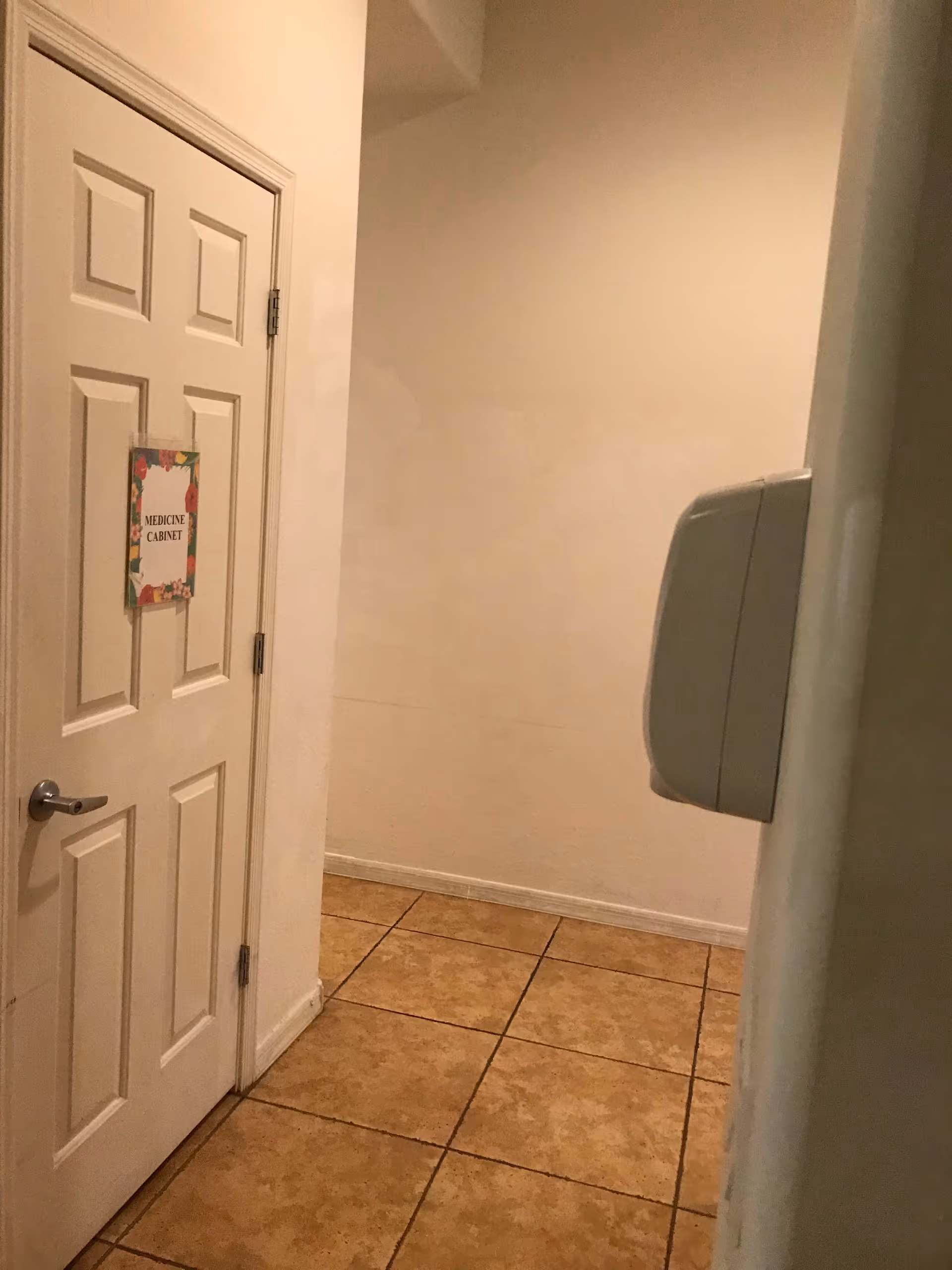 A white door labeled Medicine Cabinet stands in a beige tiled hallway with a wall-mounted dispenser.