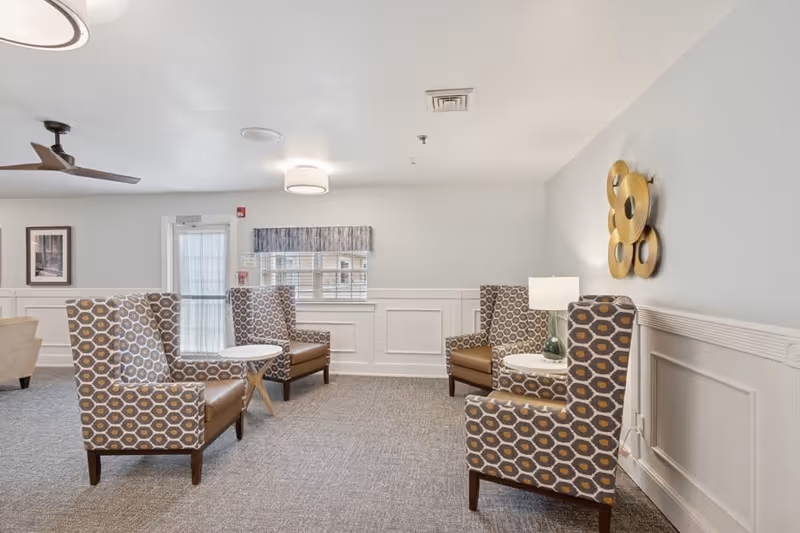 A bright and clean sitting area with four patterned armchairs arranged around two small white round tables. The room has light gray walls with white wainscoting, a ceiling fan, a window with a valance, and a decorative gold wall art piece. There is a table lamp on one of the tables providing soft lighting.