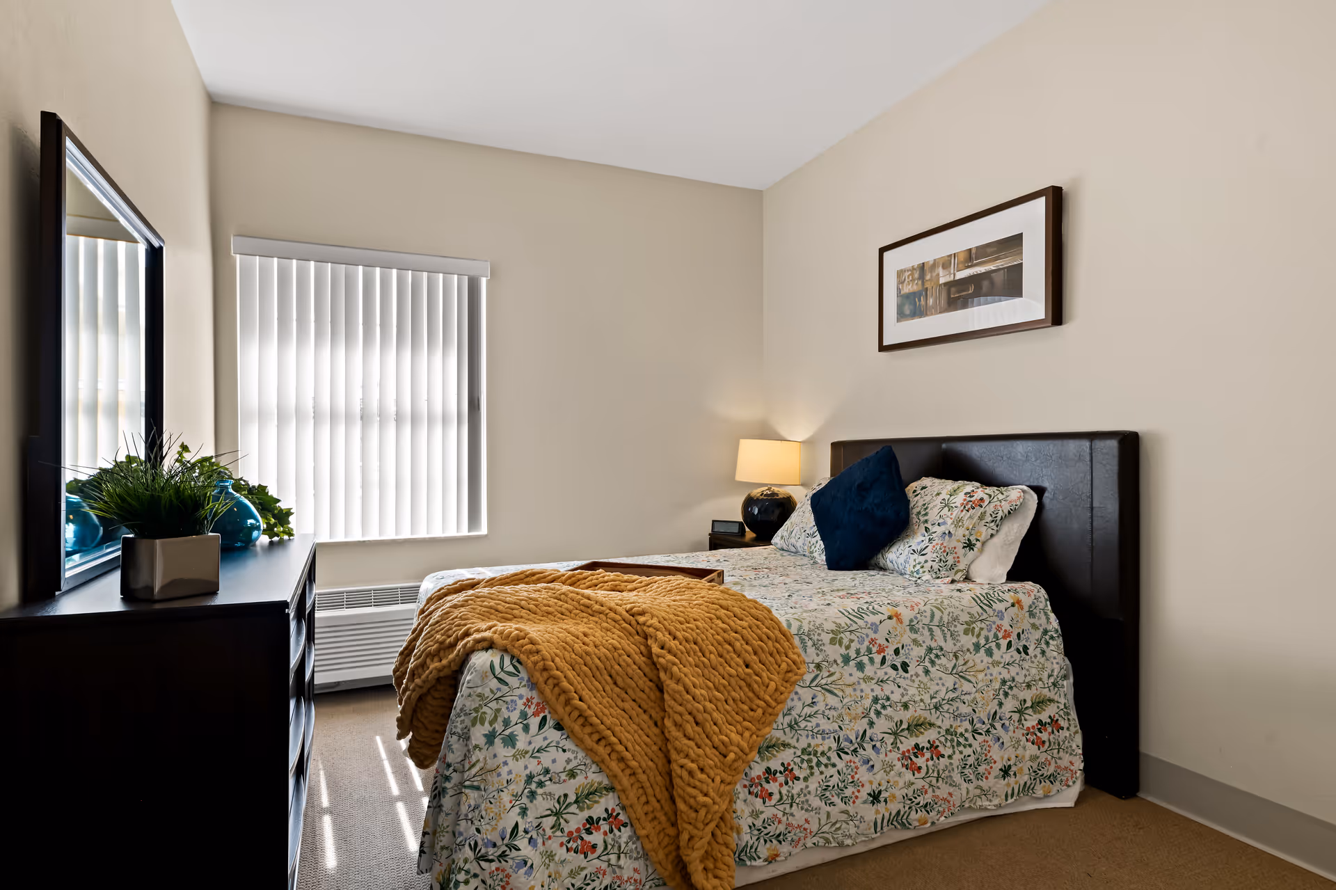 A cozy bedroom with a bed covered in floral bedding and a mustard yellow knitted throw blanket. There are two pillows on the bed, one dark blue and one floral. A nightstand with a lamp and an alarm clock is beside the bed. A dresser with a large mirror and decorative plants is on the left side of the room. The window has vertical blinds partially closed, allowing light to filter in.