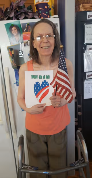 An elderly woman standing indoors in front of a refrigerator and a bulletin board, holding a small American flag and a paper with a heart decorated in stars and stripes and the text 'Happy 4th of July'. She is smiling and using a walker.