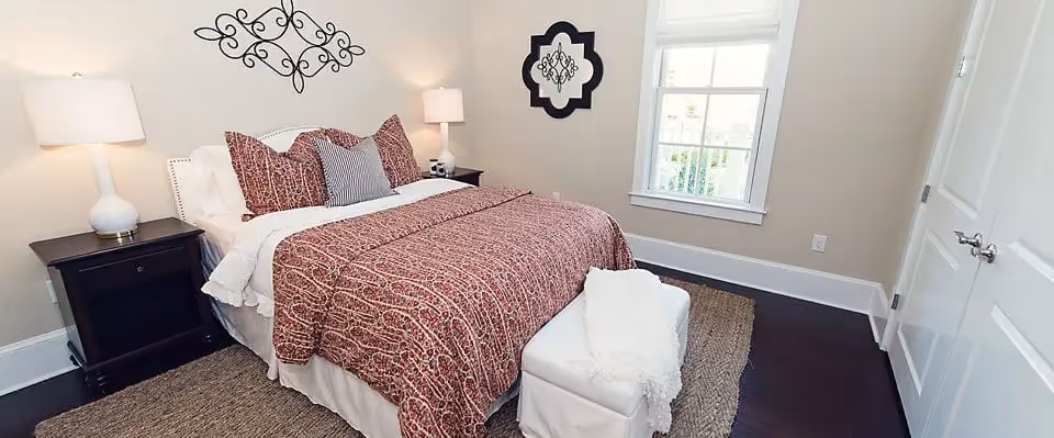 A bedroom with a neatly made bed featuring a red patterned comforter and multiple pillows. There are two dark wooden nightstands on either side of the bed, each with a white lamp. The room has beige walls, a window with white blinds, a decorative wall hanging above the bed, and a white cushioned bench at the foot of the bed on a woven rug.