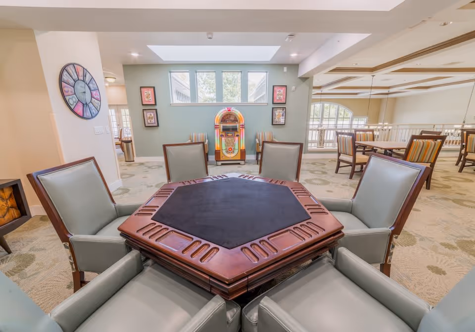 Bright communal lounge with a hexagonal game table surrounded by chairs, a colorful jukebox against a green wall, and dining tables in the background.