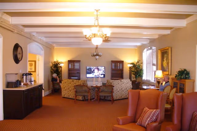 A cozy living room area in a senior living facility with beige walls and a carpeted floor. The room features a television mounted on the far wall, flanked by two wooden cabinets. There are two floral-patterned sofas and two wooden chairs arranged around a small table in front of the TV. Two elderly individuals are seated separately on either side of the room. The ceiling has exposed beams and two chandeliers. There is a sideboard with coffee supplies on the left and a window with blinds and a lamp on the right. The room is warmly lit with table lamps and wall sconces.