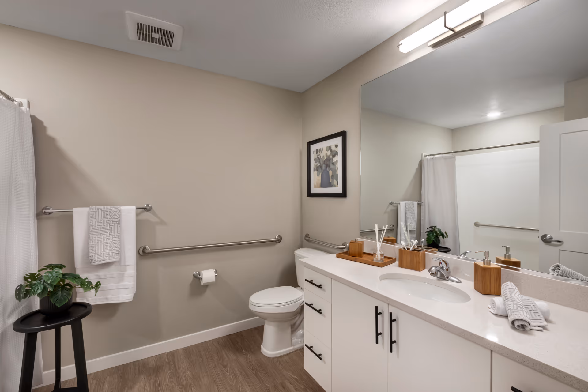 A clean and modern bathroom with beige walls and wood-look flooring. The room features a white toilet, a white vanity with a sink, and a large mirror above the vanity. There are handrails installed near the toilet and along the wall. A towel rack with two white towels is mounted on the wall next to the shower area, which has a white curtain. A small black stool with a green potted plant is placed near the towel rack. The countertop has decorative items including a soap dispenser and a reed diffuser. A framed picture hangs on the wall above the toilet.