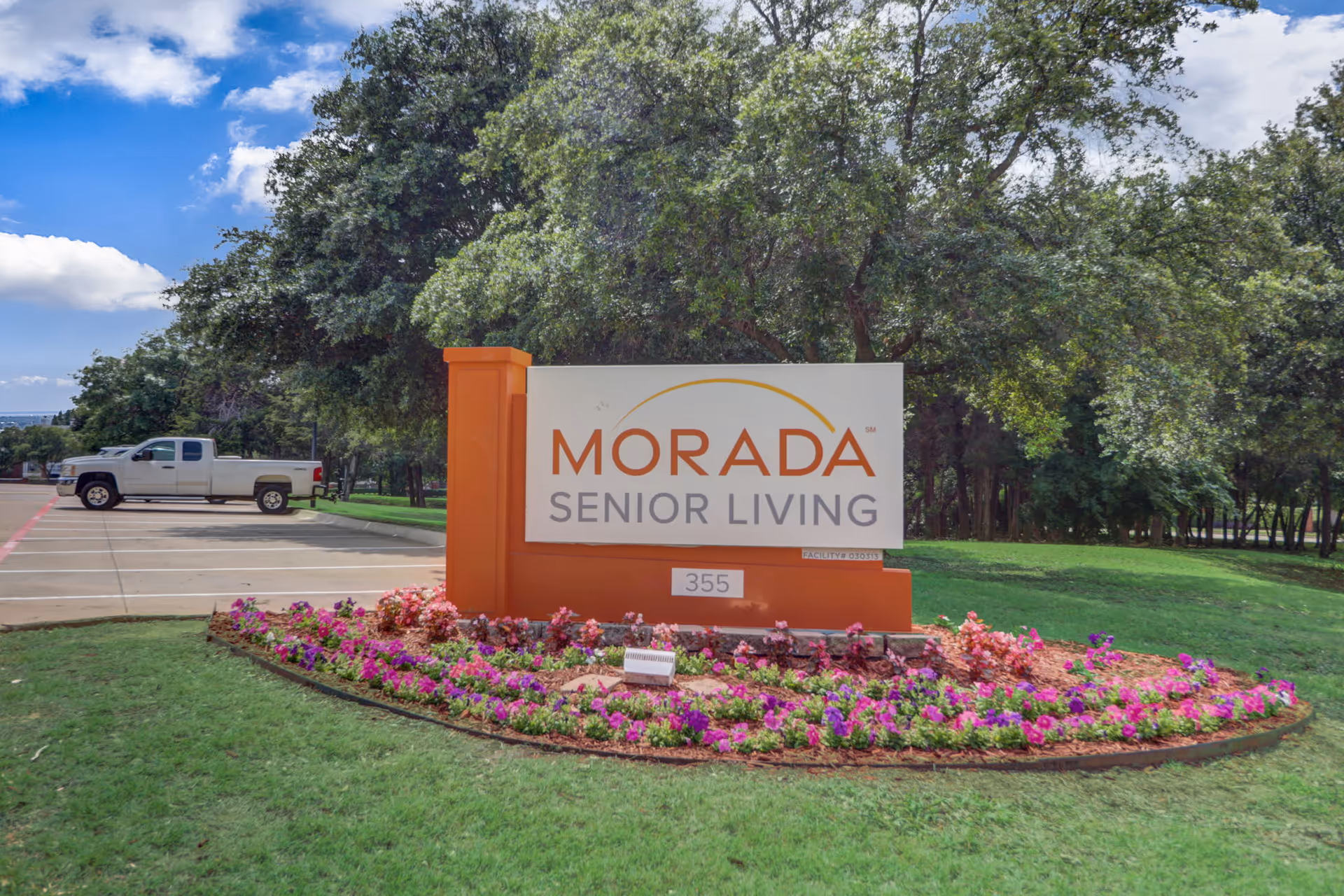 Outdoor view of the Morada Senior Living facility sign surrounded by a flower bed with pink and purple flowers, green grass, and large trees in the background under a partly cloudy blue sky. A white pickup truck is parked on the left side in a parking lot.
