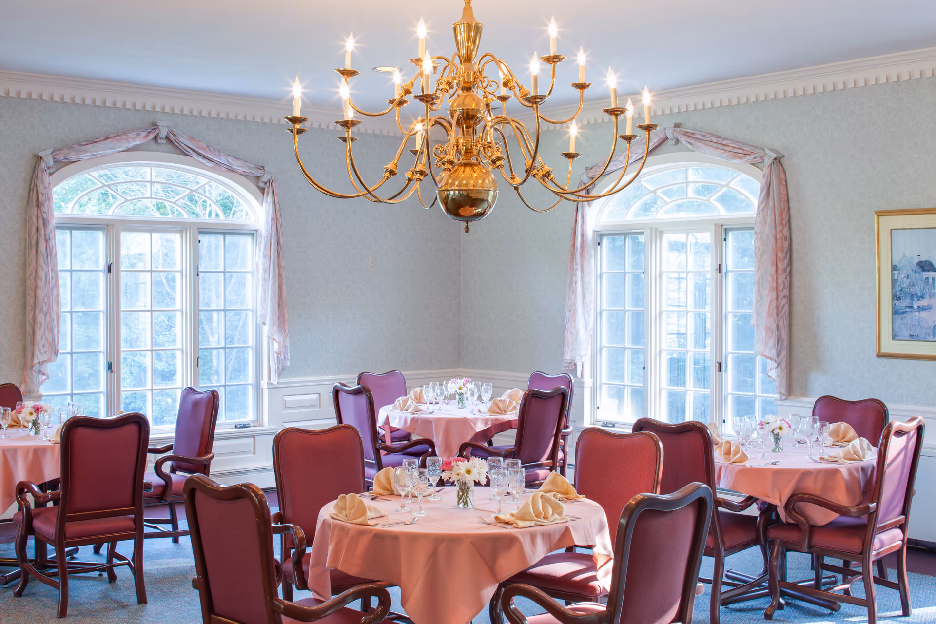 A dining room with round tables covered in pink tablecloths, each set with folded napkins, glassware, and small floral centerpieces. The room features large arched windows with sheer curtains, a decorative chandelier hanging from the ceiling, and upholstered chairs with wooden frames around each table.