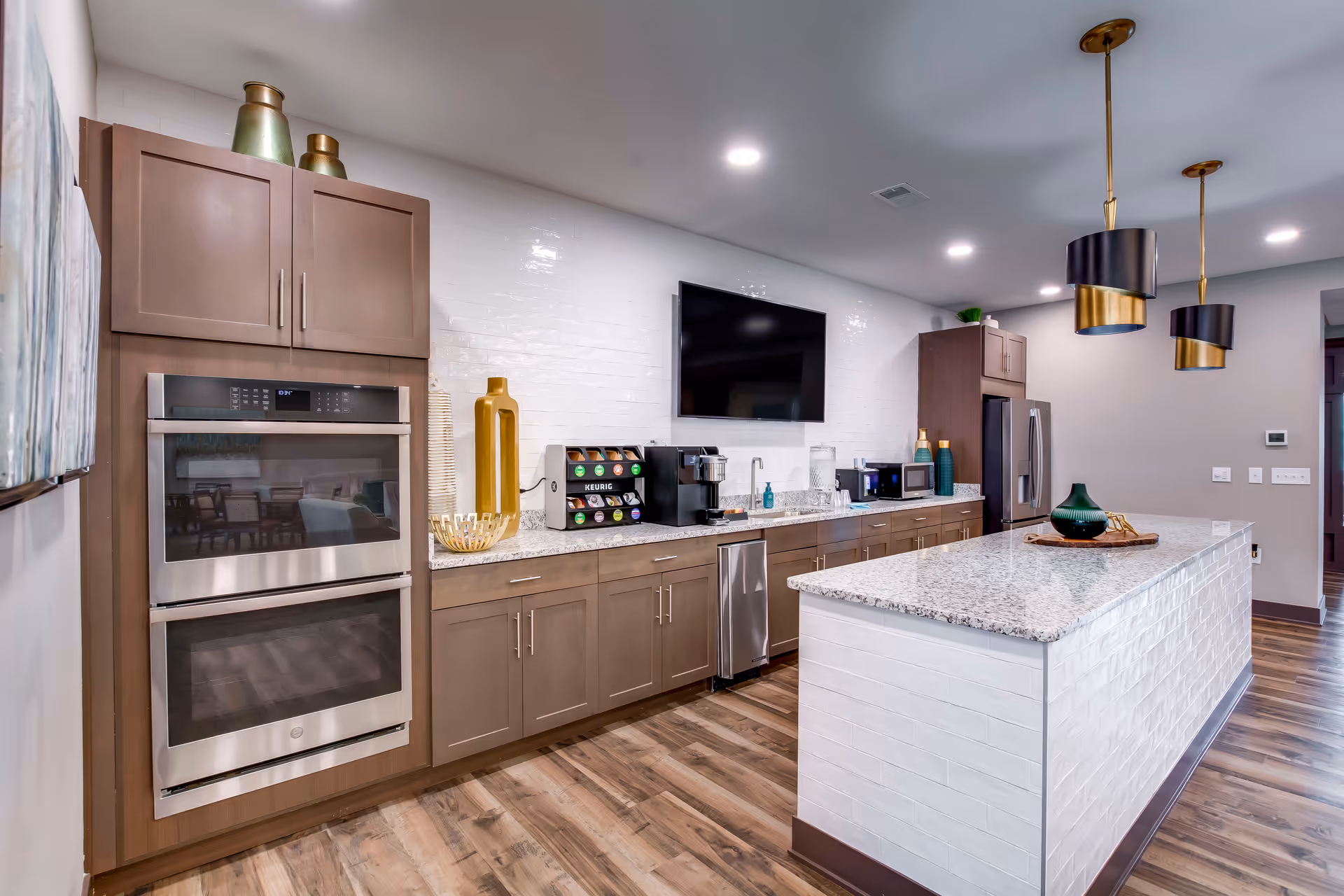 Bright modern community kitchen with a large granite island, pendant lights, stainless appliances, and a coffee station.