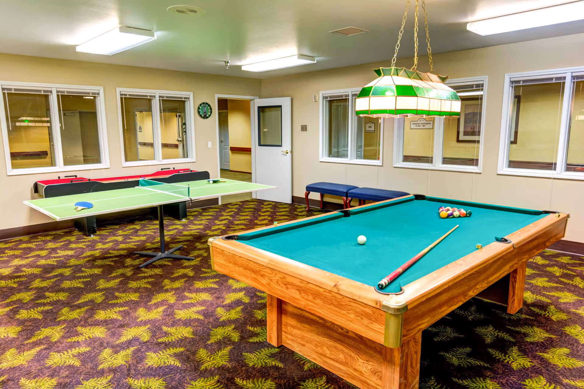 A recreational room with a pool table, a ping pong table, and a foosball table. The pool table has a cue stick and billiard balls arranged on it. The room has patterned carpet, beige walls, several windows, a green and white hanging light fixture above the pool table, and a bench along one wall.
