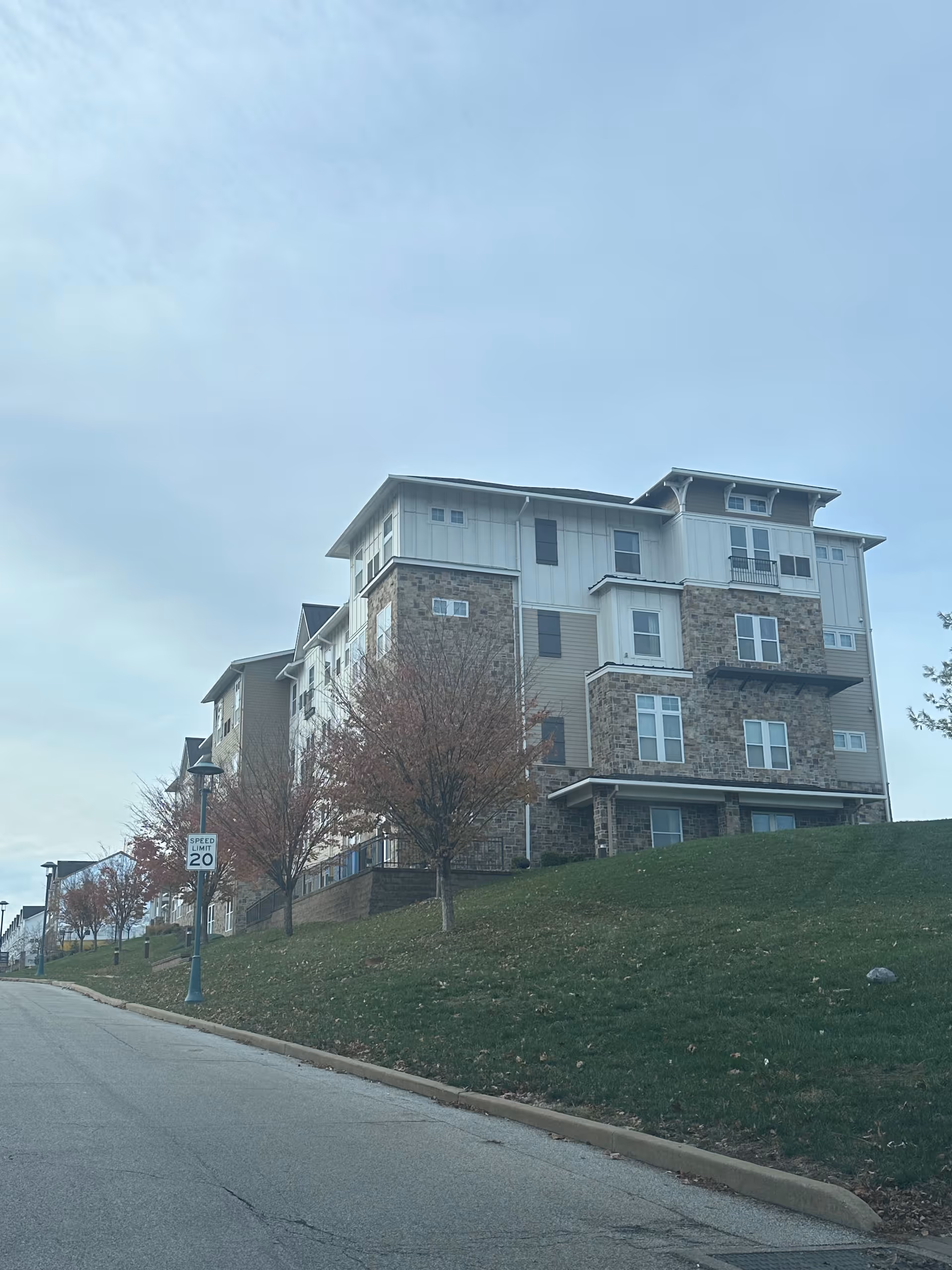 Multi-story residential building with a stone-and-siding facade on a grassy slope beside a street.
