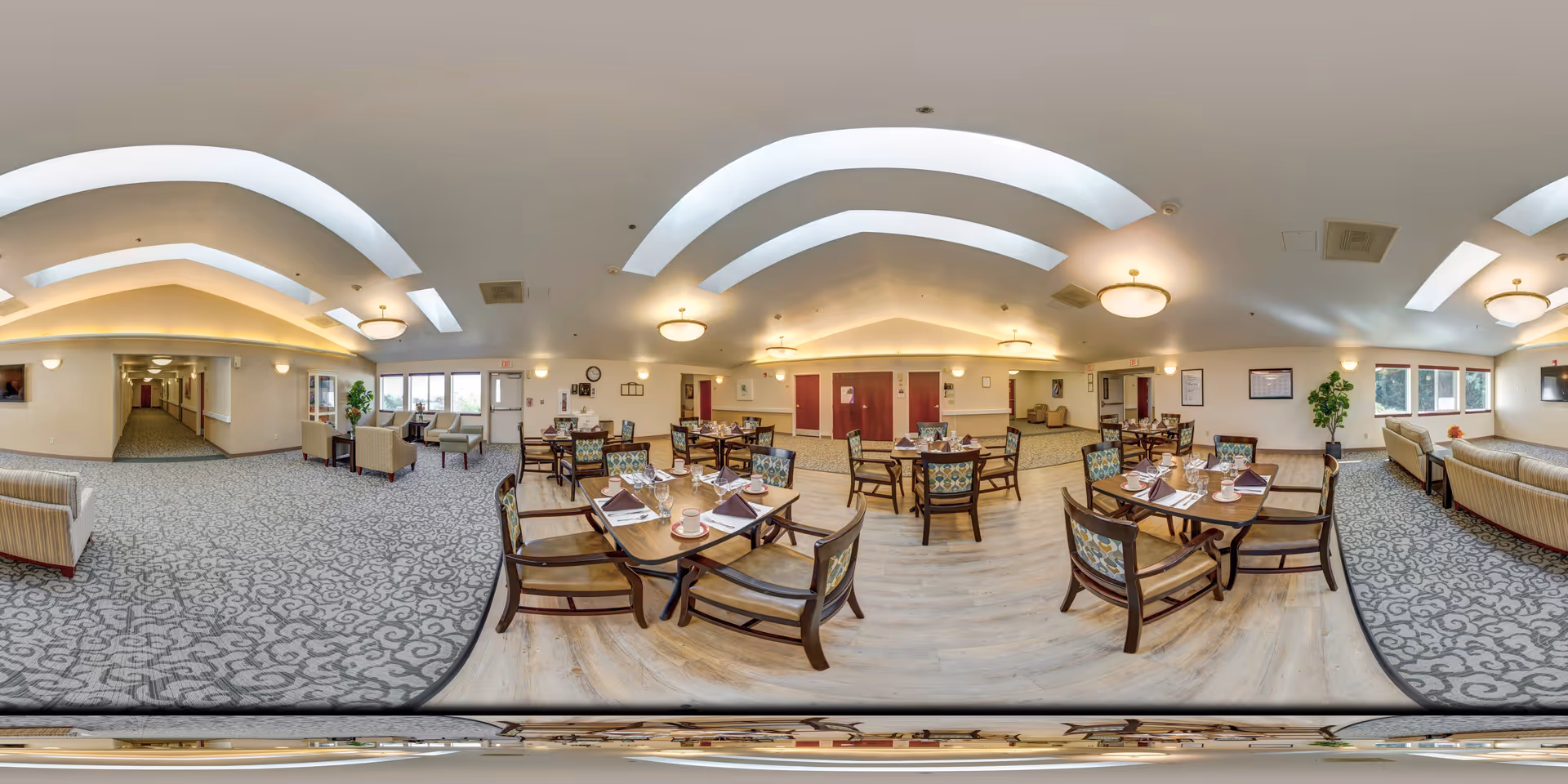 A spacious, well-lit dining room in Monarch Gardens Memory Care with several wooden tables and chairs arranged neatly. Each table is set with placemats, napkins, cups, and utensils. The room features patterned carpet and wood flooring, comfortable seating areas with sofas and armchairs, large windows allowing natural light, and ceiling lights with modern fixtures. The walls are light-colored with framed pictures and plants adding a touch of greenery.