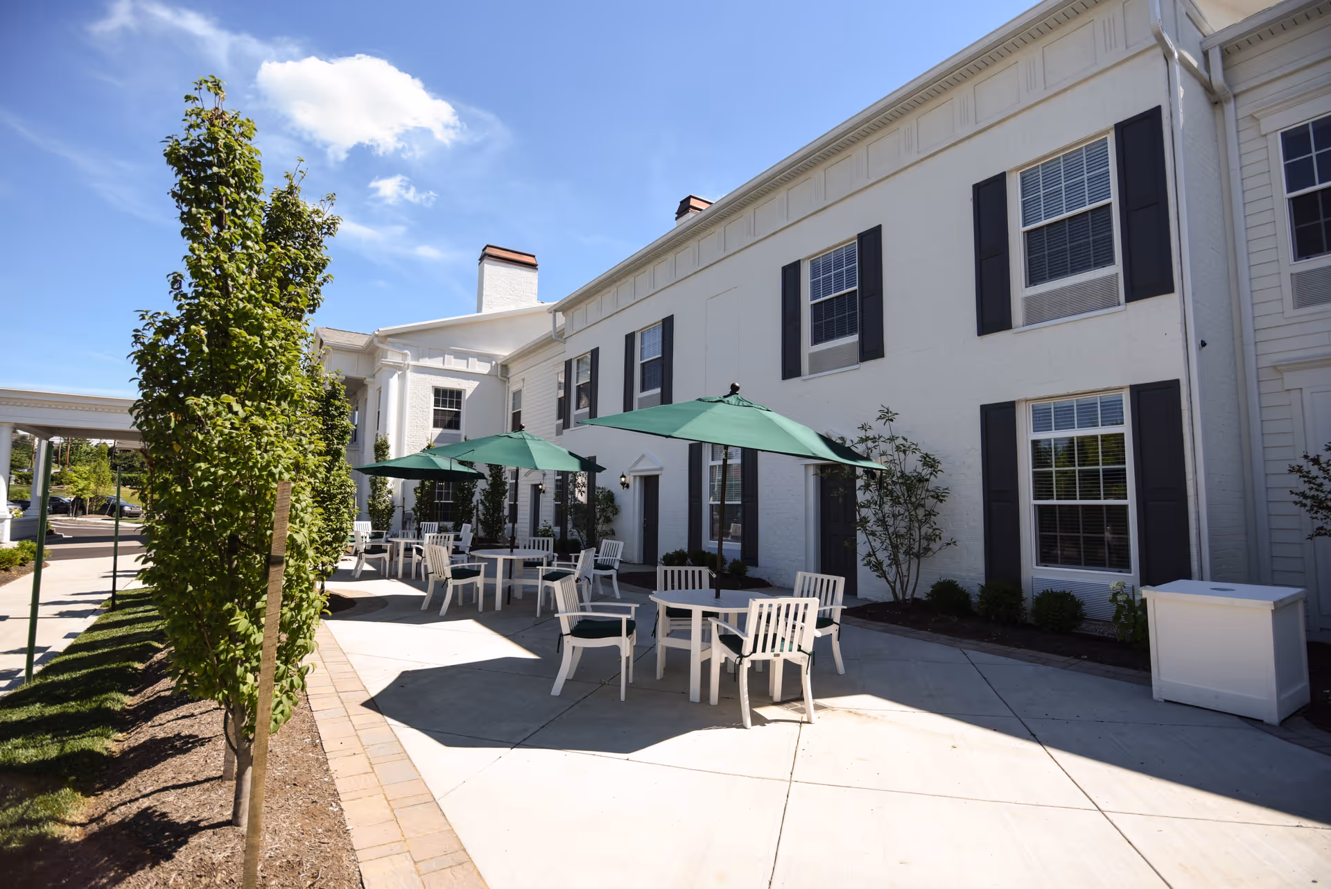 Outdoor patio area at Highgrove at Tates Creek with white tables and chairs under green umbrellas, adjacent to a white building with black window shutters. There are small trees and shrubs planted along the walkway under a clear blue sky.