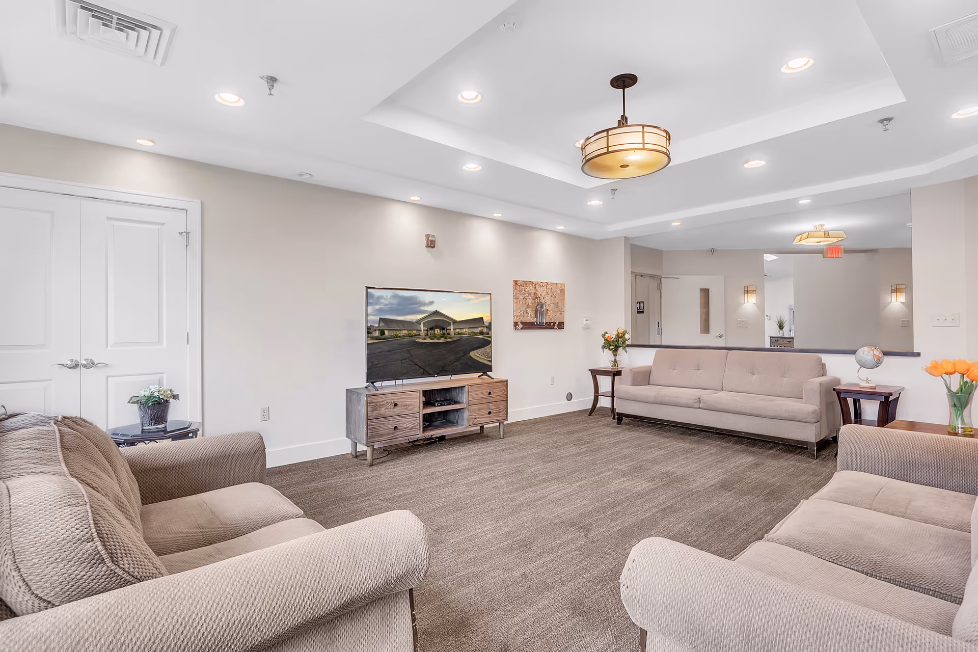 Bright, spacious common living area with multiple beige sofas, a TV on a wooden stand, side tables with flowers, and recessed ceiling lights.
