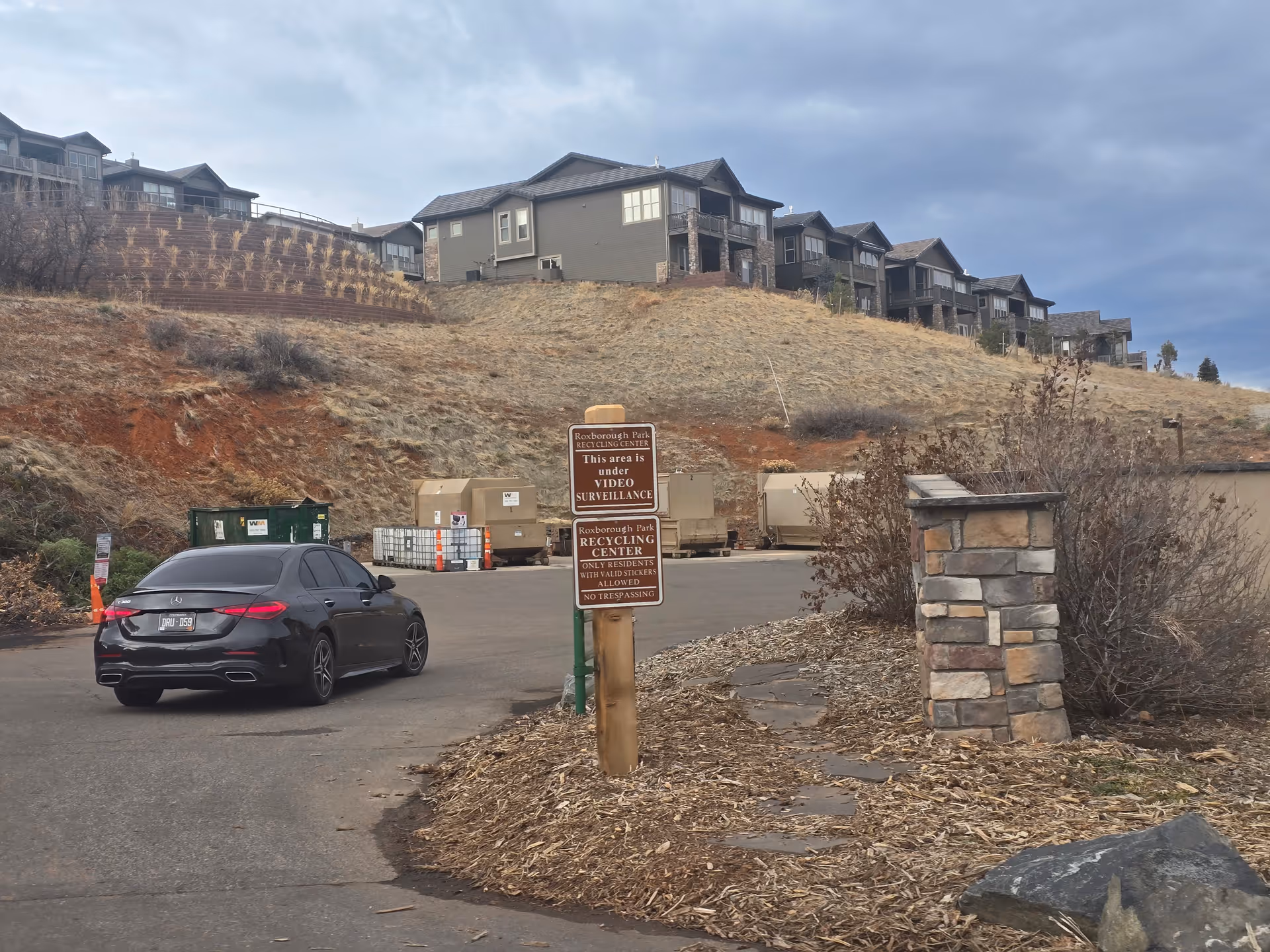 A black car driving on a paved road towards a recycling center with several large dumpsters. The area is surrounded by dry grass and bushes, with houses on a hill in the background under a cloudy sky. A signpost near the road reads 'Roxborough Park Recycling Center' with rules about video surveillance and resident access.