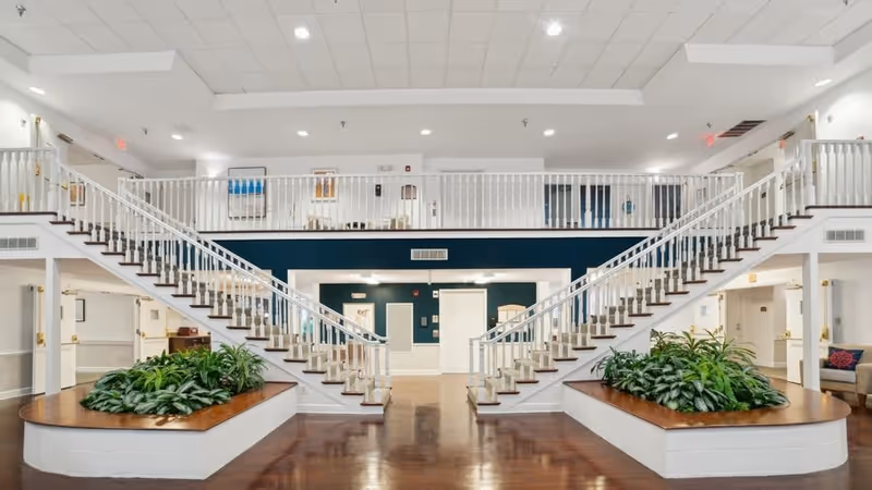 Spacious interior of a senior living facility featuring a large open area with two symmetrical staircases leading to an upper balcony. The staircases flank two large planters filled with green plants. The walls are white with dark blue accents, and the floor is polished wood. The ceiling has recessed lighting, and there are doors and seating areas visible in the background.