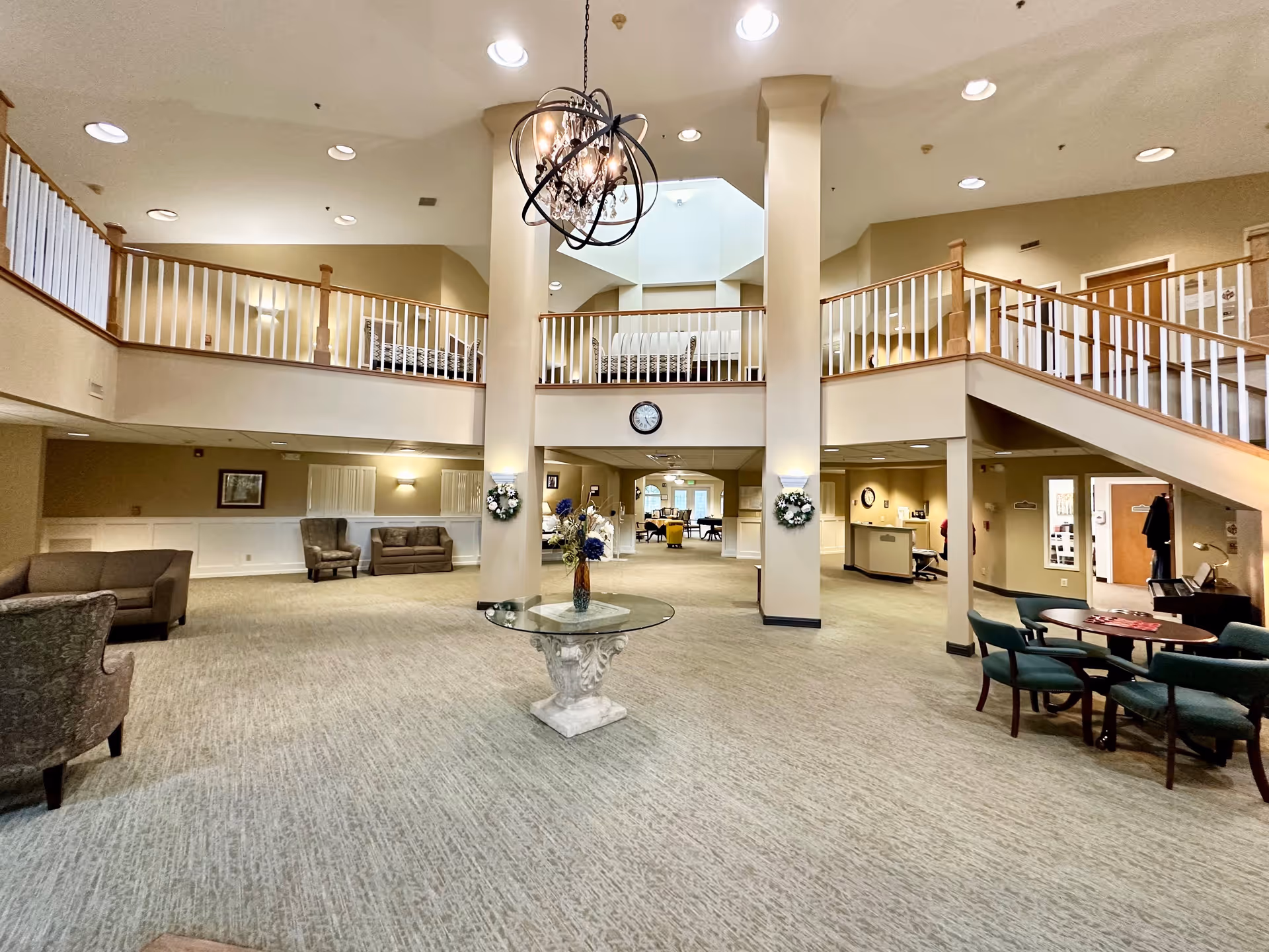 Spacious senior living community lobby with high ceilings, a central glass table with a floral arrangement, multiple seating areas with sofas and chairs, a staircase leading to an upper balcony, and warm lighting fixtures including a chandelier.