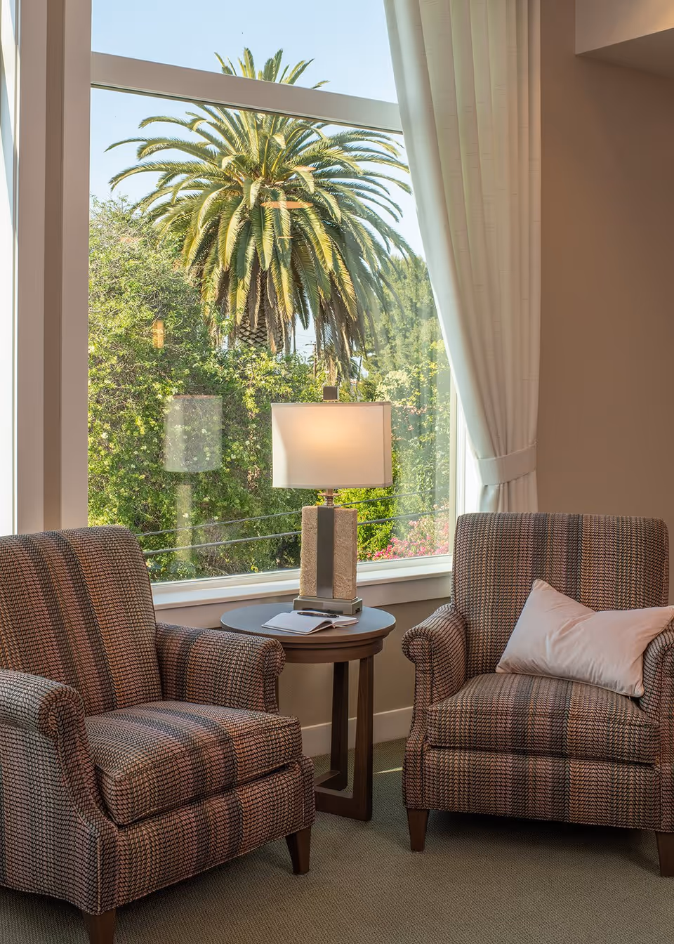 Two patterned armchairs flank a round side table with a lamp in front of a large window overlooking a palm tree and garden.