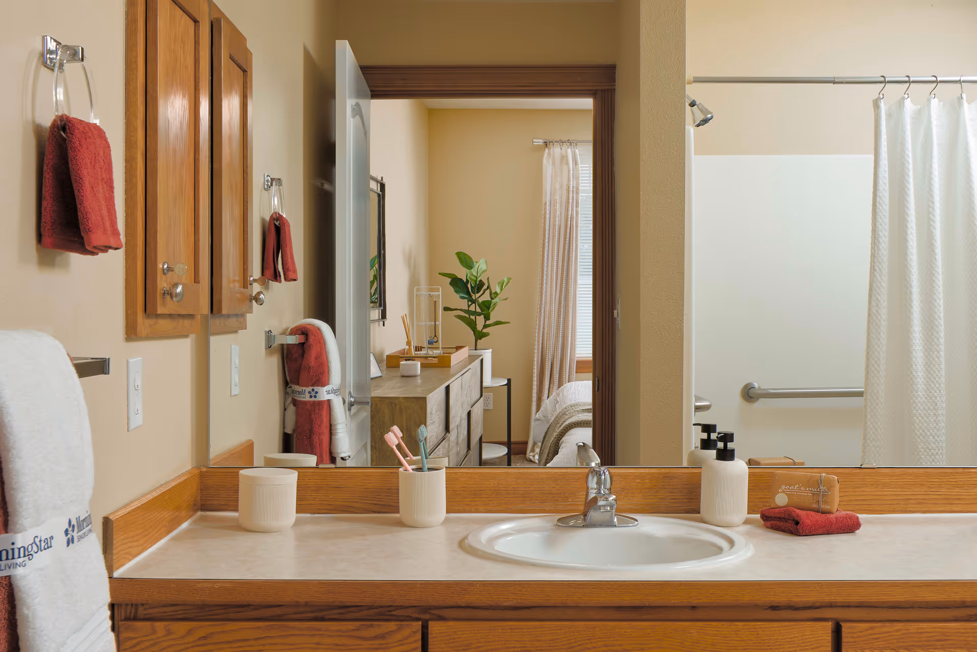 Bathroom sink area with a large mirror reflecting a bedroom. The countertop has a white sink, toothbrush holder with toothbrushes, soap dispenser, and a small towel. Red towels hang on the wall and are folded on the counter. The bathroom has beige walls and wooden cabinets. A shower with a white curtain and grab bar is visible in the reflection.