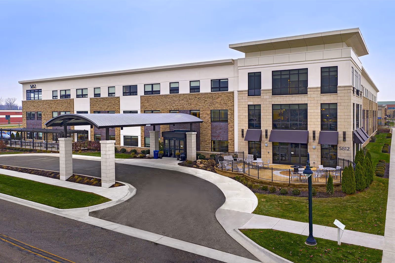 Exterior view of a modern three-story senior living facility building with a covered entrance driveway, large windows, and a small fenced patio area with outdoor seating. The building has a mix of beige stone and brick facade with a well-maintained lawn and sidewalk surrounding it.