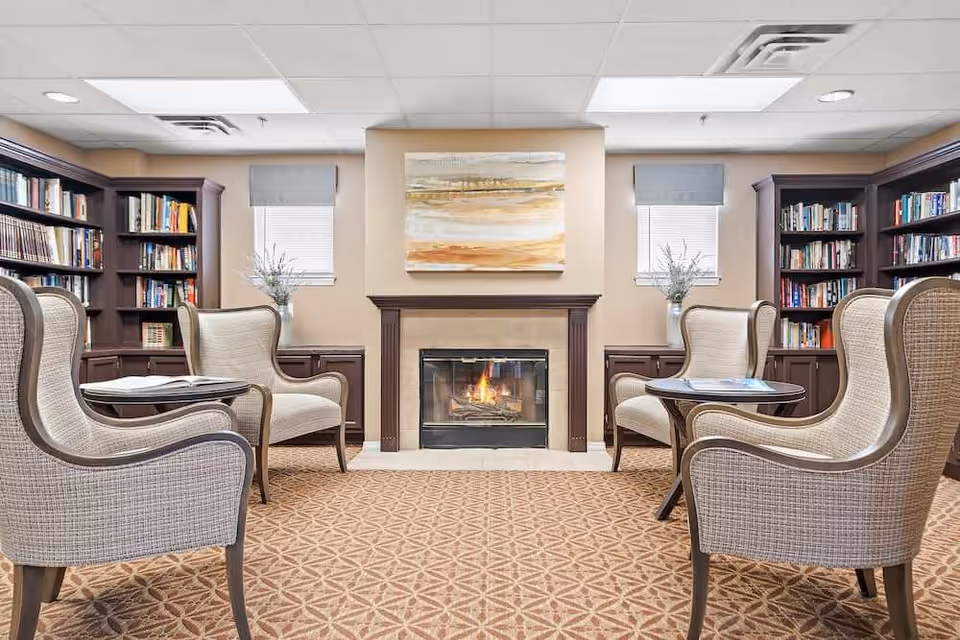 A cozy reading room with four upholstered armchairs arranged around two small round tables, a lit fireplace in the center, bookshelves filled with books on both sides, two windows with blinds and vases with flowers on the cabinets beneath them, and a painting above the fireplace.