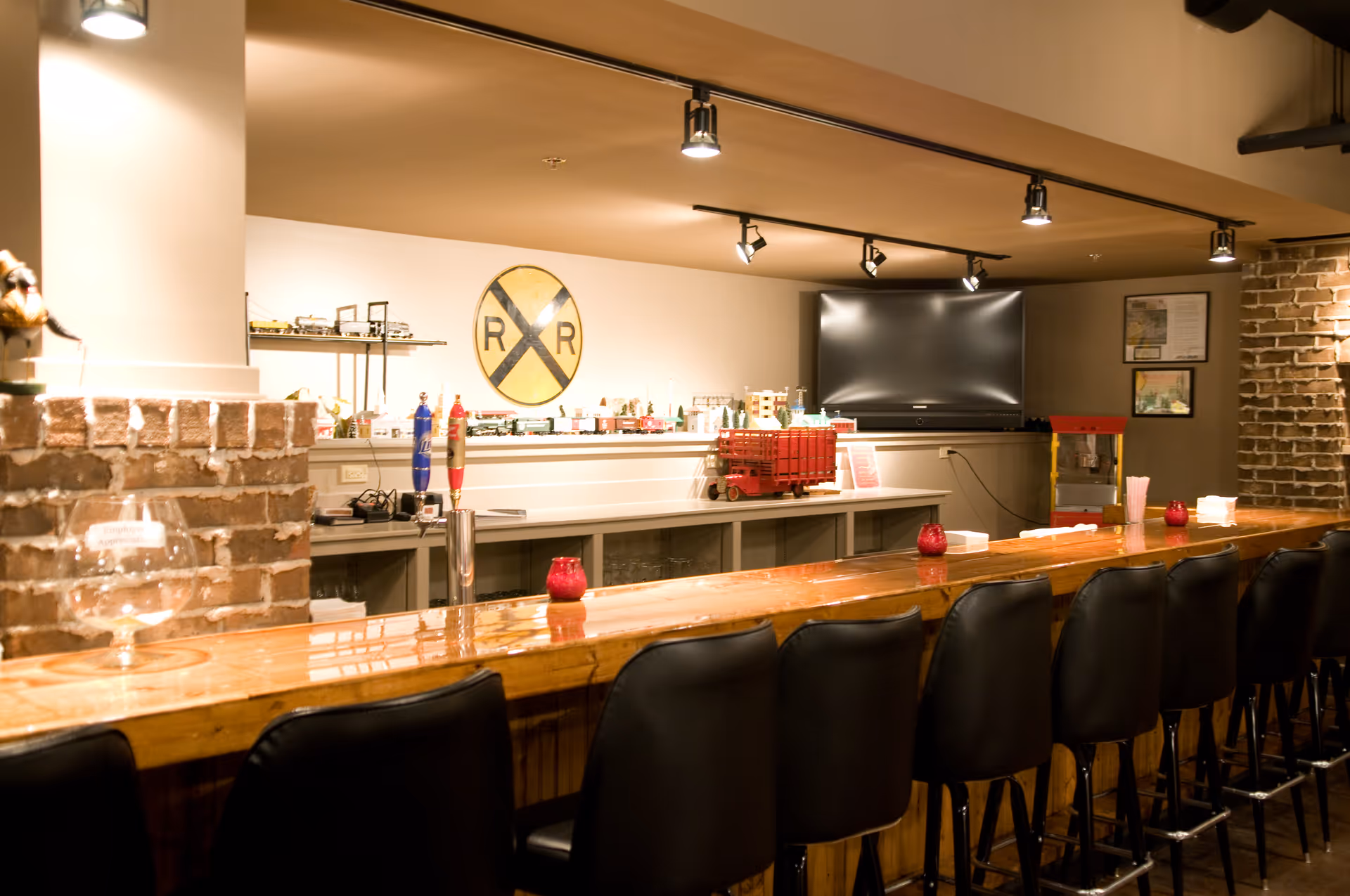 Interior view of a bar area with a long wooden counter and black bar stools. Behind the counter, there is a shelf with model trains and a large railroad crossing sign on the wall. A flat-screen TV is mounted on the back wall, and there is a popcorn machine to the right. The space has exposed brick walls and track lighting on the ceiling.