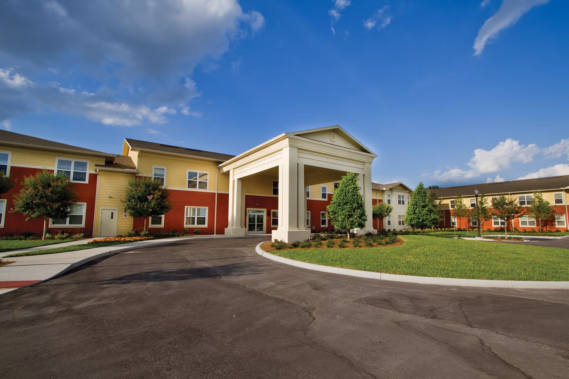 Front entrance of a two-story senior living facility with a covered portico, red and yellow siding, landscaped lawn and driveway under a blue sky.