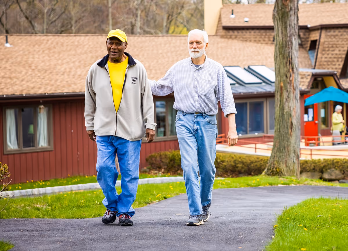 Two elderly men walking together on a paved path outside a senior living facility with buildings and greenery in the background.