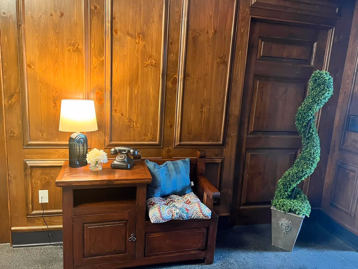 Wood-paneled interior nook with a wooden bench and side table holding a lamp, vintage telephone, small flowers, and a spiral topiary in a pot.