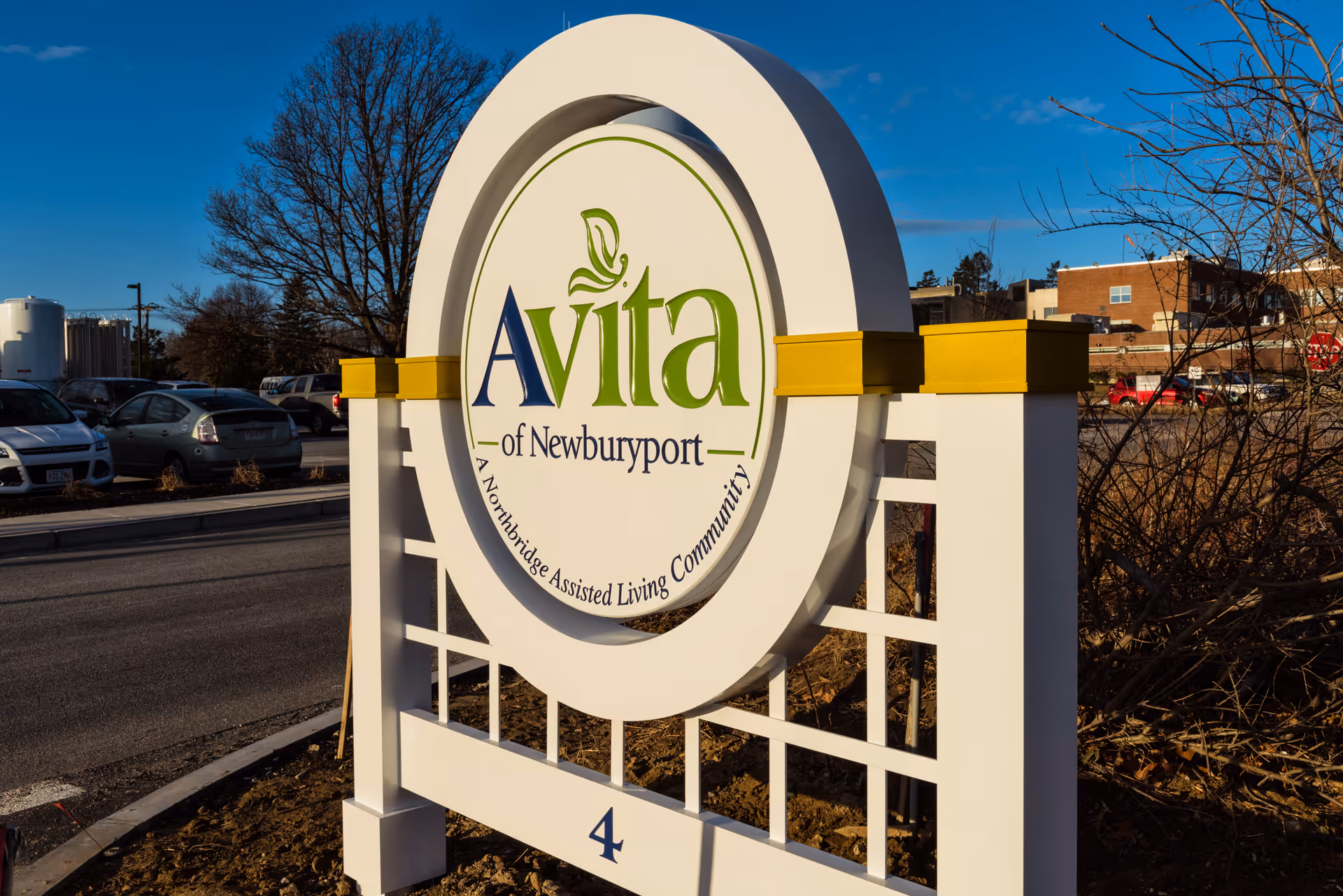 Outdoor sign for Avita of Newburyport, a Northbridge Assisted Living Community, located near a parking lot with cars and some buildings in the background under a clear blue sky.