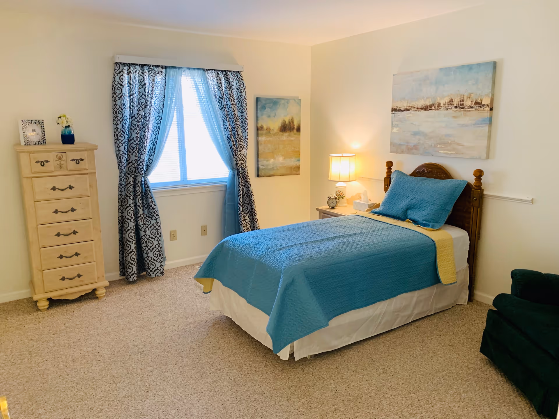 A cozy bedroom with a single bed covered in a blue and yellow quilt. Next to the bed is a wooden nightstand with a lamp, alarm clock, and tissue box. There is a wooden dresser with decorative items on top, and a window with patterned blue curtains letting in natural light. Two paintings hang on the walls, and a dark green armchair is partially visible.