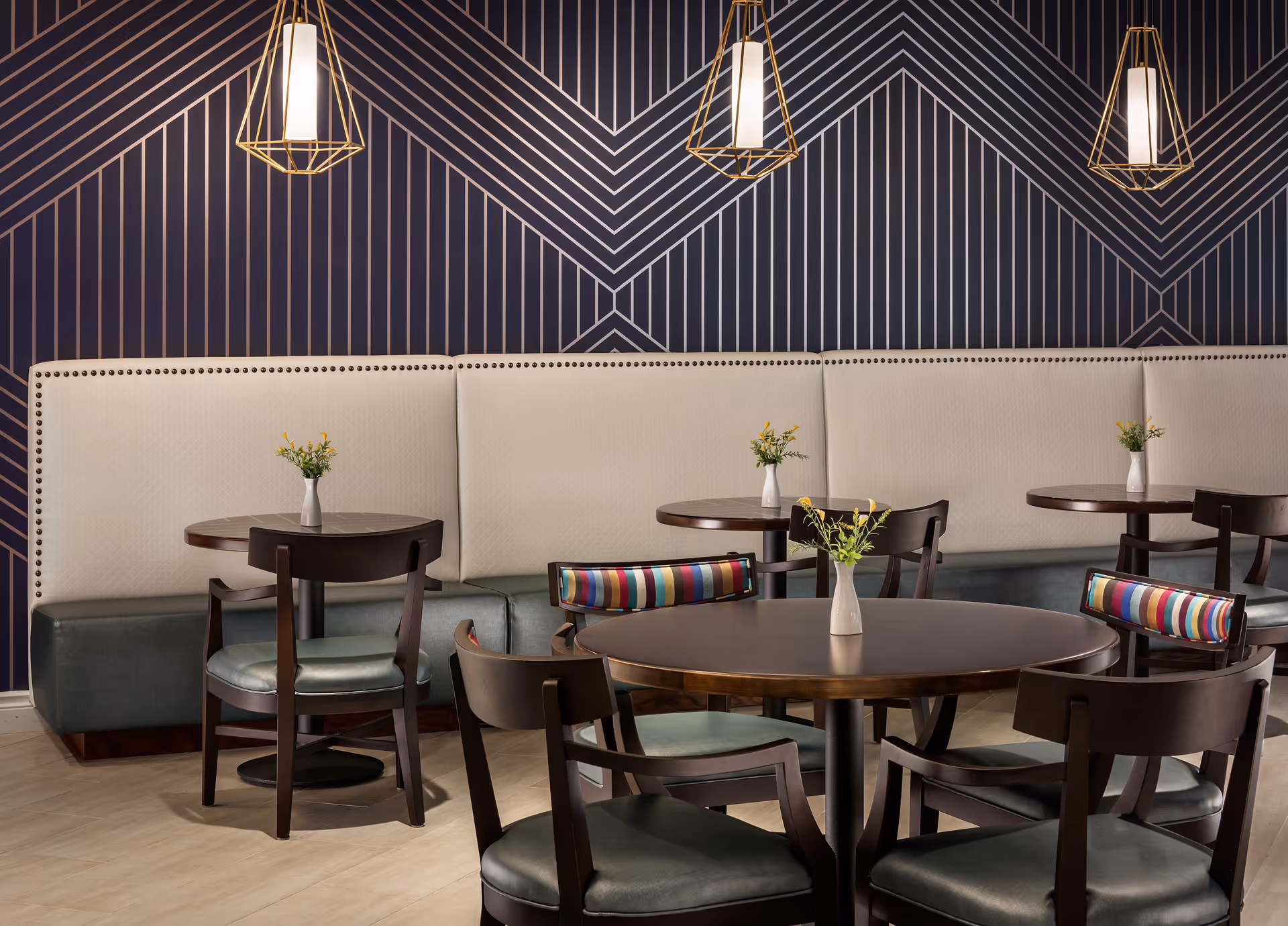 Stylish dining area with round wooden tables, dark chairs, a beige banquette against a geometric patterned wall, pendant lights, and small vases of flowers.