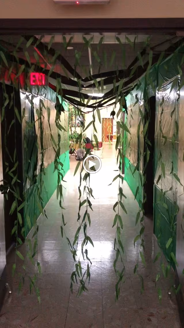 An indoor hallway decorated with green hanging streamers like vines leading toward a lit room with an EXIT sign.