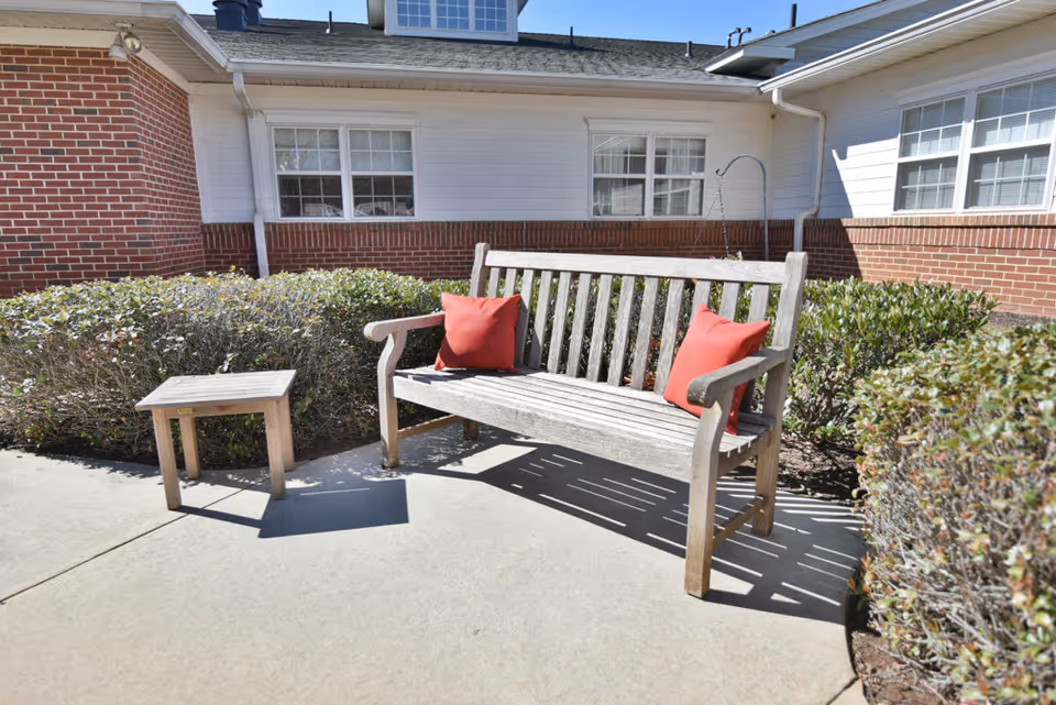 Outdoor patio area at Oaks at Chanticleer with a wooden bench featuring two red cushions and a small wooden side table, surrounded by trimmed bushes and the exterior walls of the building with windows.