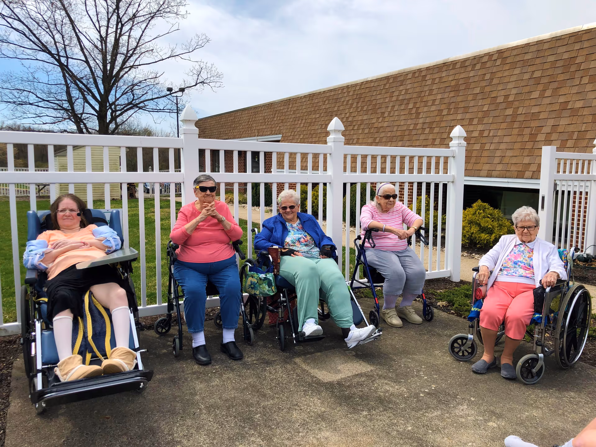 Five elderly women sitting outside in wheelchairs and a walker near a white fence with a building in the background. They are enjoying the outdoor weather on a paved area with some grass and trees visible.