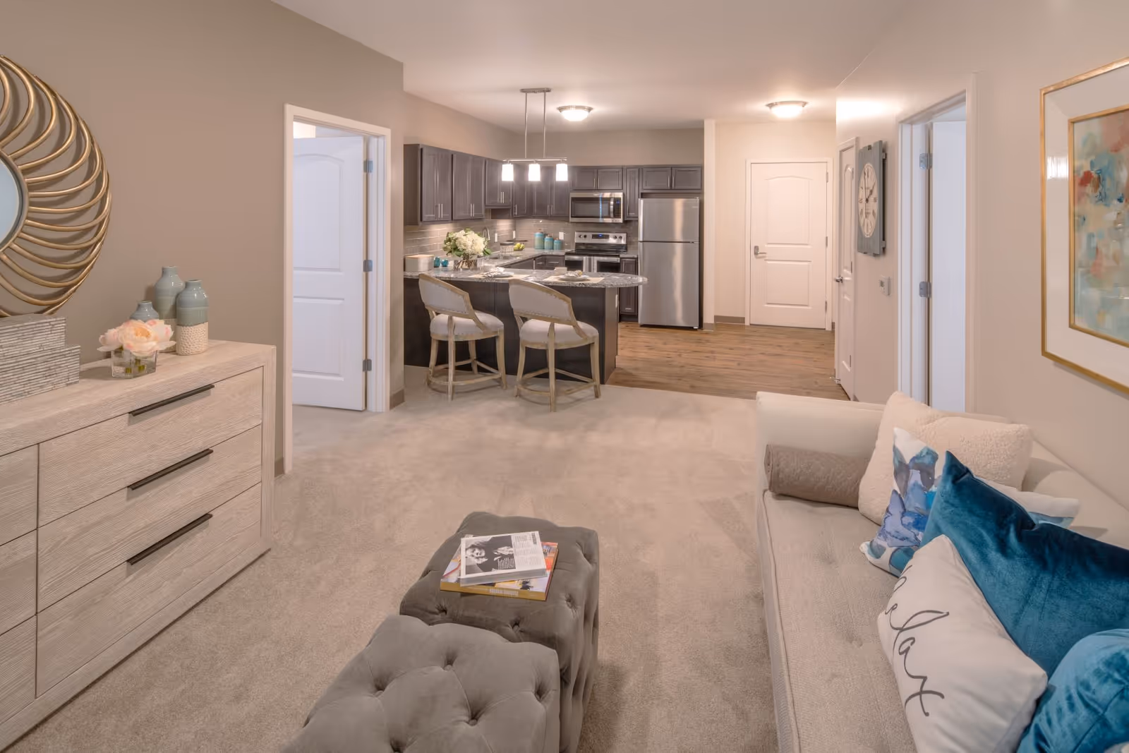 Open living room leading to a modern kitchen with an island, sofa, ottoman, and dresser.