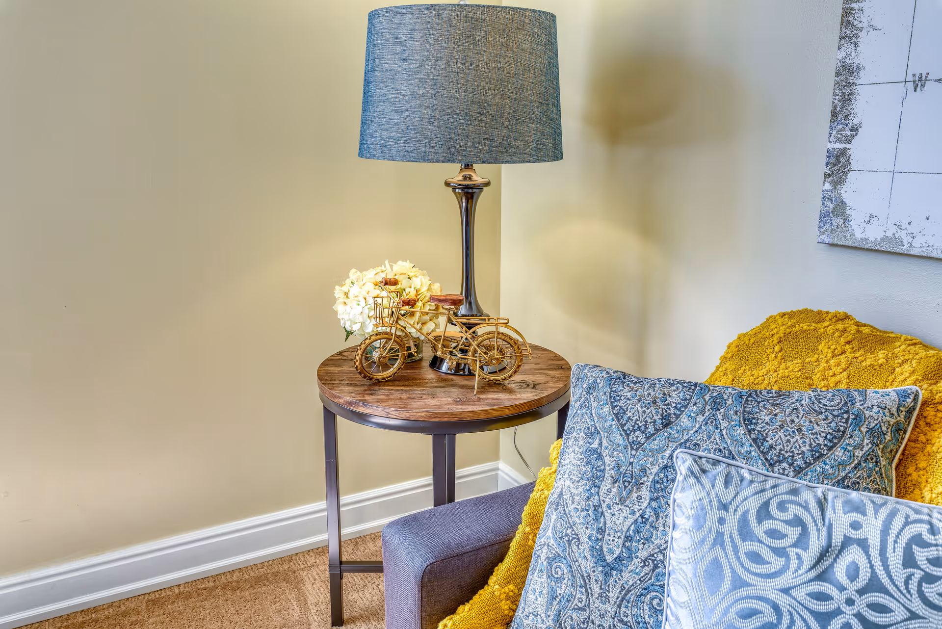 Side table with a lamp, decorative bicycle and flowers next to a couch with patterned pillows and a yellow throw.