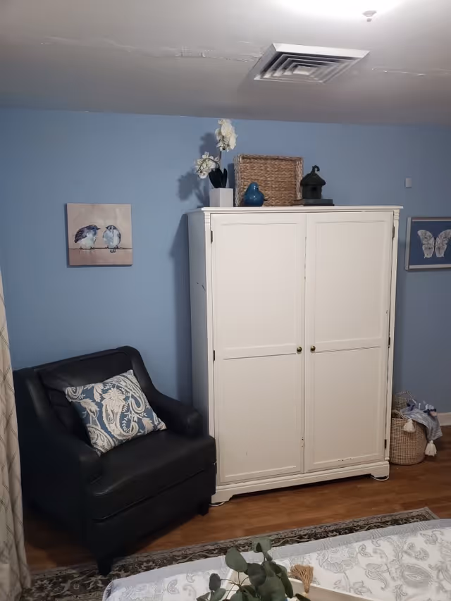 A cozy corner of a room with light blue walls featuring a black armchair with a patterned cushion, a white cabinet topped with decorative items including a basket, a blue bird figurine, and a small plant. There are framed pictures of birds and butterflies on the walls, a basket with clothes in the corner, and part of a bed with a floral bedspread visible in the foreground.